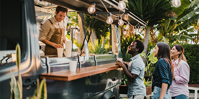 several customers being served at a food truck in a tropical area, palm trees and greenery behind them