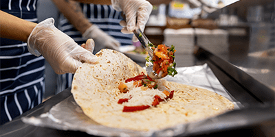 gloved hands folding a burrito in a restaurant, placing a final meat topping over them