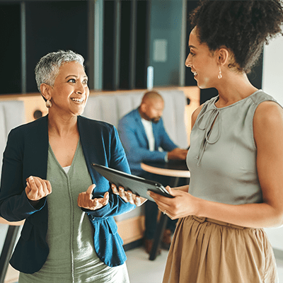 an elderly woman with short gray hair in business atire seems to be offering advice or explaining something to a younger woman who has a clipboard or tablet to take notes