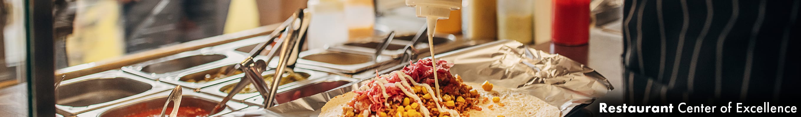 cook placing sauce on a burrito, standing in front of a grid of ingredients where customers can see and choose their ingredients for a burrito