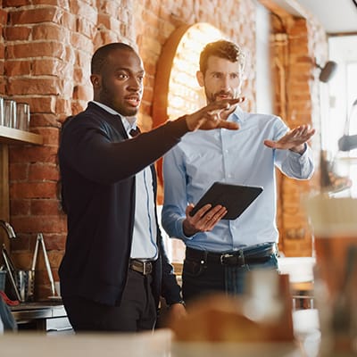 two men discussing decor options in a restaurant behind a bar, an old brick wall behind them