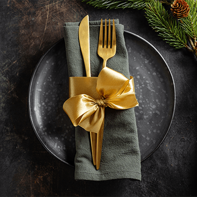 A dark table, charcoal napkin folded on a charcoal plate, gold silverware wrapped on top with a gold ribbon knotted into a bow