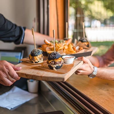 small gourmet burgers on wooden platter being handed to pair of customers