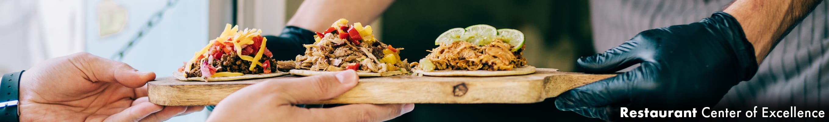 a gloved employee handing off a wooden platter with three tacos on it, to the customer