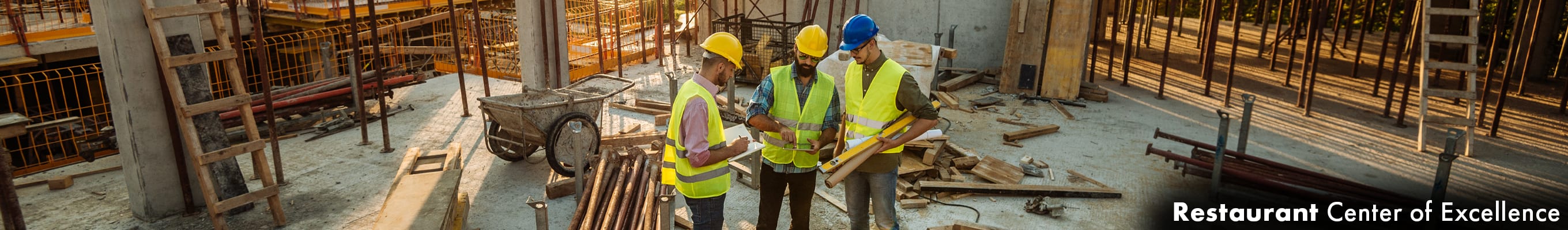 three construction workers standing and discussing plans while referencing a tablet, amidst a large construction project site