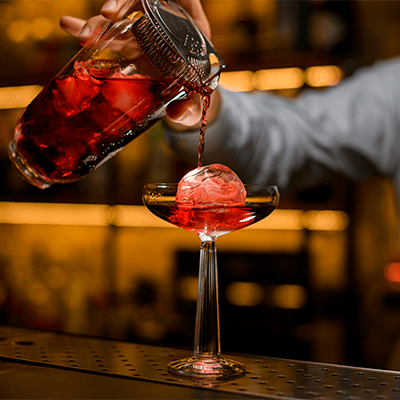 someone pouring a iced red drink into a tall glass with a sphere of ice that breaks the stream of liquid into the glass