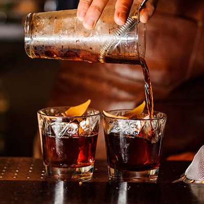a skilled bartender pouring a dark drink into two short glasses with orange peals sitting over the ice