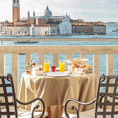 small dining table covered in horderves and narrow orange juice glasses, overlooking beautiful waters and European architecture in the distance