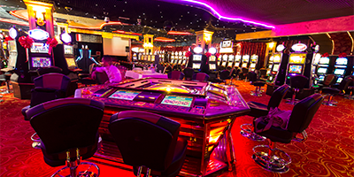 A casino with a red carpet, a game table empty of players nearby, and in the background there's a wall of flashing colorful slot machines