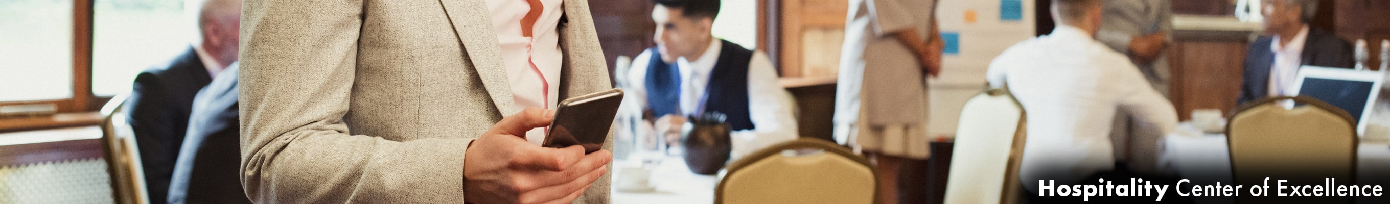 a man on his phone, behind him it appears there is a conference going on, circular tables, people in suits, a woman standing in front of a display