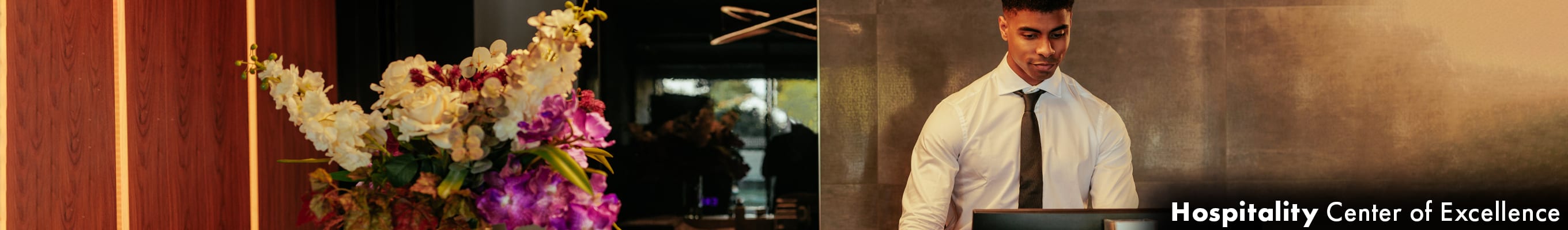 front desk of a hotel, a large vase of flowers to the left, a man in a suit on a monitor to the right