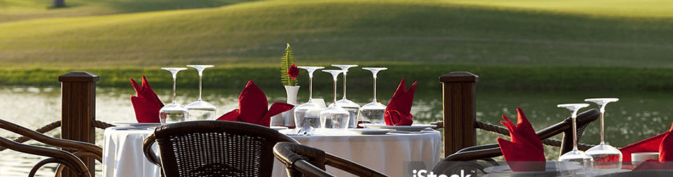 An elegant outdoor dining setup overlooking a serene lake, featuring a table with fine glassware and folded red napkins, ready for a high-end golf, social, or country club event.