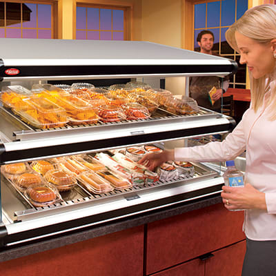 FOH Equipment A woman selecting food items from a heated display case in a store, with a man in the background.