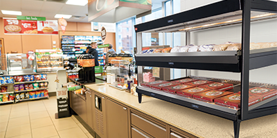 Equipment & Supplies An interior view of a convenience store with a pizza warmer on the counter, a customer at the checkout, and shelves stocked with various items in the background.