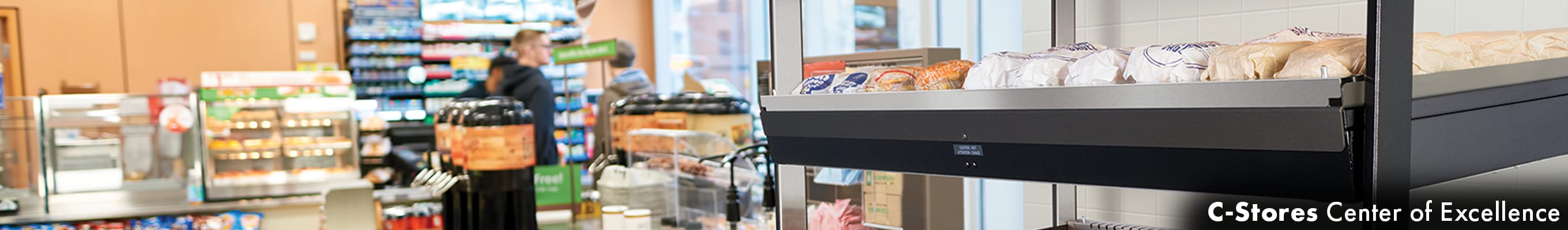 A panoramic interior view of a convenience store with a coffee station and food warmer in the foreground and a customer at the checkout in the background.