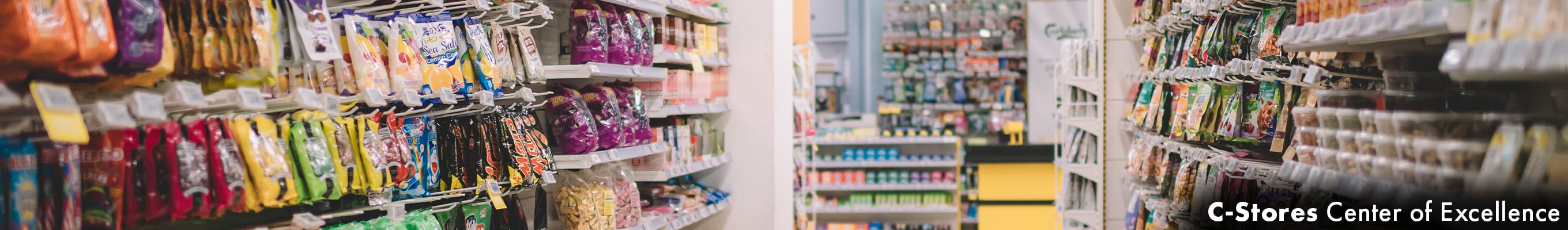 convenience store aisle, brightly colored packaged items to the left and right along the shelves, a checkout counter in the distance
